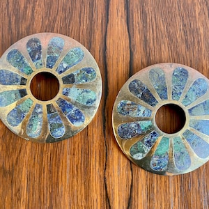 May include: Two decorative, circular objects with a central hole. The objects have a gold-colored metal frame with petal-shaped inlays of blue and green stone. The background is a dark wood surface.