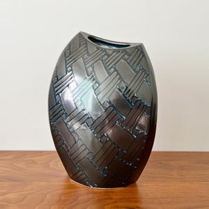 May include: A ceramic vase with an oval silhouette and a dark metallic gray finish. The surface features a raised, repeating woven basket-weave pattern with subtle blue accents visible within the recessed areas of the textured design.