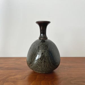 May include: A ceramic vase with a bulbous, rounded body and a narrow, flared neck. The base of the vase features a mottled, dark gray and black speckled glaze, while the neck is finished in a smooth, solid dark brown glaze.