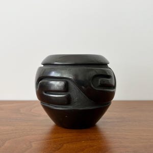 May include: A small, rounded black pottery vessel with a smooth, polished finish. The exterior features carved, recessed geometric patterns that wrap around the midsection, creating a textured contrast against the otherwise sleek, dark surface.
