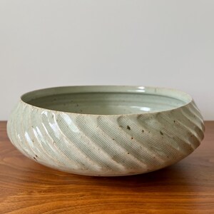 May include: A light green ceramic bowl with a textured, ridged exterior. The bowl has a wide, shallow shape and sits on a wooden surface. The interior of the bowl is a lighter shade of green.