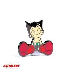 May include: A black and white enamel pin featuring Astro Boy, a classic Japanese manga character, sitting with his arms crossed and wearing a green belt. The pin is red and has a shiny finish.