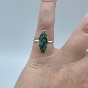 May include: A silver ring featuring an oval-shaped abalone shell inlay in shades of green and blue. The ring is displayed on a finger. The abalone shell has a distinctive swirling pattern, set against a white backdrop.