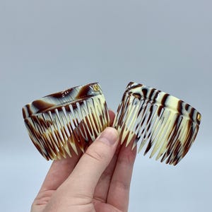 Könnte beinhalten: Zwei Haarkämme mit Schildpattmuster. Die Kämme haben eine gebogene Form mit feinen Zähnen und einem marmorierten Design in Braun-, Creme- und Beigetönen. Jeder Kamm hat einen kleinen goldfarbenen Akzent.