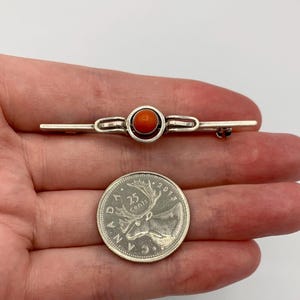 May include: A silver-toned bar brooch featuring a central circular setting with a smooth, round orange stone. The brooch is positioned above a Canadian 25-cent coin, which measures approximately 2.38 cm in diameter, for scale reference.