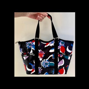 May include: Black tote bag with a floral pattern in red, white, blue, and purple. The bag has black handles and a zip closure. The bag has two exterior pockets on each side.