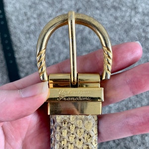 May include: A gold-coloured belt buckle with the engraved name "Valentino Franchini" is attached to a snakeskin-patterned belt. The buckle has a twisted rope design on the sides. The belt is held by a hand.