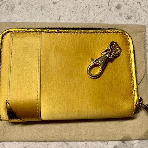 May include: A gold wallet with a zip and a gold-coloured clasp. The rectangular wallet has a smooth texture and sits on a beige fabric. The wallet is approximately 12 cm long.