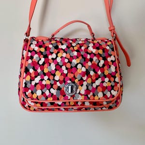 May include: A Vera Bradley crossbody bag featuring a quilted pattern of colourful dots in pink, orange, yellow, grey, and white. The bag has a coral-coloured trim, handle, and shoulder strap. A silver clasp secures the flap.