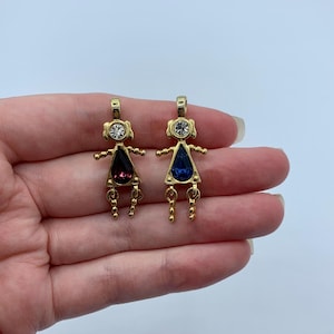 May include: Two gold-coloured pendant earrings shaped like stick figures. Each figure has a teardrop-shaped gemstone, one purple and one blue. The figures have small arms and dangling legs. A clear crystal is set in the head of each figure.