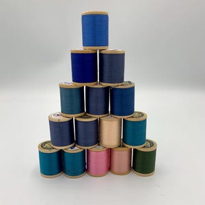 May include: A pyramid stack of fifteen wooden spools of sewing thread in various shades of blue, teal, pink, and beige. Each spool features a cylindrical wooden core wrapped with coloured thread, arranged in five rows with a single spool on top.