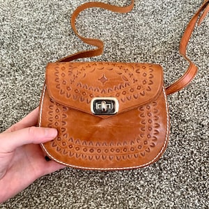 May include: A tan leather crossbody bag with an embossed pattern. The bag has a flap closure with a silver clasp and a long strap. The edges are detailed with white stitching. The bag is approximately 20cm wide.