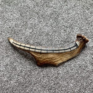 May include: A curved brown deer antler featuring an inlaid cribbage board track with black and white markings. The board includes a series of small drilled holes for scoring and the text WELLS printed in black ink near the base of the antler.