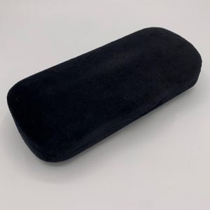 May include: A rectangular, black, velvet-textured hard-shell case with rounded corners. The exterior surface has a soft, matte finish and a uniform dark black color. The case is closed, featuring a simple, clean design with no visible hardware.