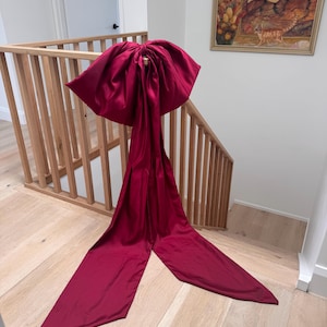May include: A large, deep red satin bow with long, flowing tails is draped over a wooden staircase railing. The bow's voluminous shape and rich colour create a striking visual contrast against the light wood and white walls.