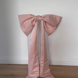 May include: A large, blush pink bow made of a silky fabric. The bow is mounted on a clear acrylic stand, with long, flowing tails. The bow is centered in the frame, against a white wall and wooden floor.