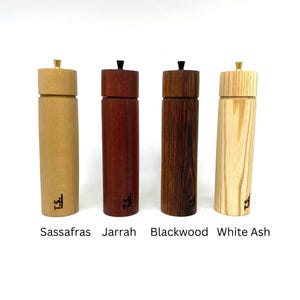 Könnte beinhalten: Vier Pfeffermühlen aus Holz in verschiedenen Holzarten: Sassafras, Jarrah, Blackwood und Esche. Jede Mühle hat ein Messing-Oberteil und einen schwarzen Knopf.