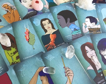 May include: A collection of rectangular oracle cards featuring diverse illustrations of people, nature, and animals against a teal background. The cards include text such as I AM POWERFUL, I AM LIMITLESS, and VULNERABILITY INSPIRES, accented by clear quartz crystals.