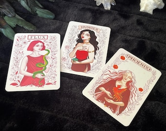 May include: Three tarot cards featuring illustrations of women. The cards are labelled "Flux", "Hunger", and "Phoenix". The "Flux" card shows a woman in a red dress holding a green snake. The "Hunger" card depicts a woman in a red top. The "Phoenix" card shows a woman with a bird.