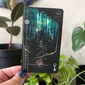 May include: A black card featuring a forest scene and a key symbol. The card has the word "DISTILL" printed vertically on the left side and the number "42" printed in the top right corner.
