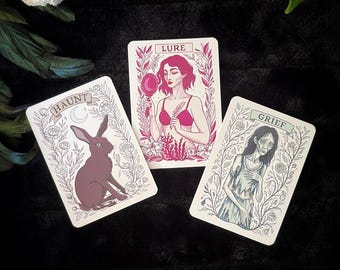 May include: Three tarot cards with illustrations and text. The "Haunt" card features a rabbit, the "Lure" card shows a woman with a mirror, and the "Grief" card depicts a woman. Each card has floral borders and a cream background.