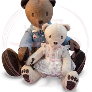 Op de afbeelding: Twee pluche teddyberen. Eén is bruin en draagt een blauw jasje met een witte vlinderdas. De andere is crèmekleurig en draagt een witte jurk met roze bloemenaccenten en kanten rand. Beide hebben donkerbruine oren en poten.