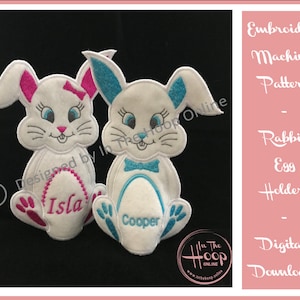 Könnte beinhalten: Stickmuster für eine Hasen-Eierhalter. Das Muster enthält zwei Hasen, einen mit rosa Akzenten und den anderen mit blauen Akzenten. Die Hasen halten Eier, auf denen "Isla" und "Cooper" steht.