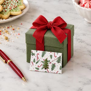 May include: A square olive green gift box tied with a wide red fabric ribbon bow, accompanied by a small envelope featuring a pattern of green pine trees and red candy canes, and a red ballpoint pen with gold-colored accents.