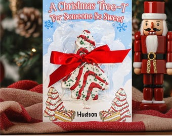 May include: A Christmas-themed gift holder featuring a Christmas tree-shaped cake with red and white stripes, tied with a red ribbon. The holder has the text "A Christmas Tree-T For Someone So Sweet" and "From: Hudson". A wooden nutcracker is in the background.