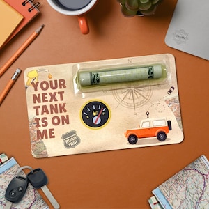 Puede incluir: Una tarjeta de regalo con el texto "YOUR NEXT TANK IS ON ME" y un gráfico de una gasolinera. La tarjeta también presenta un indicador de combustible, una brújula y un SUV naranja. Un regalo enrollado está en una funda de plástico transparente.