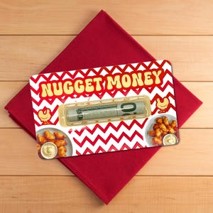 May include: A novelty gift card holder with the words "NUGGET MONEY" in yellow and red. The card features a red and white zig-zag pattern, illustrations of chicken nuggets, and a rolled-up bill. It rests on a red napkin.