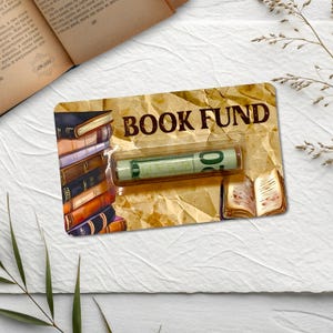 May include: A novelty gift card with the words "BOOK FUND" printed on it. The card features illustrations of books and a rolled-up bill inside a clear plastic case. The background has a vintage paper texture.