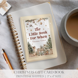 May include: A small, cream-coloured book titled "The Little Book For When Christmas Edition" with festive illustrations of Christmas trees and gifts. The book is 7.6 cm x 10.2 cm in size and rests on a white notebook.