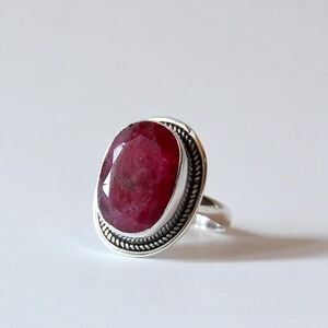 May include: A silver ring featuring a large, oval-cut ruby gemstone. The ruby is a deep red color and is set in a silver bezel with a twisted rope design around the stone. The ring band is smooth and simple.