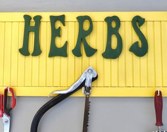 Può includere: Un cartello di legno giallo con la scritta "HERBS" in lettere verdi. Il cartello ha due ganci bianchi ed è decorato con linee verticali. Sotto il cartello ci sono attrezzi da giardinaggio, tra cui forbici e una sega.