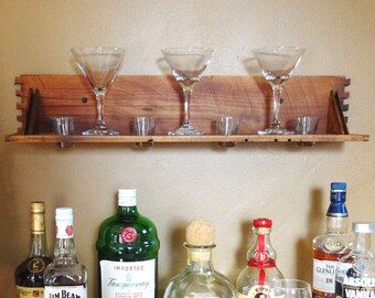 Può includere: Un'esposizione da bar con una torre Eiffel in metallo piena di tappi, una mensola in legno con bicchieri da martini e varie bottiglie di liquore. Include Jim Beam Bourbon, gin Tanqueray e vodka Absolut Vanilla. L'allestimento suggerisce un bar domestico.