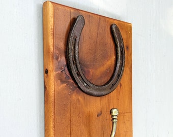 Può includere: Appendiabiti da parete rustico con un ferro di cavallo invecchiato e un gancio in ottone. Il ferro di cavallo è montato su una tavola di legno rettangolare con venature visibili. Il gancio è posizionato sotto.