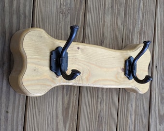 Può includere: Un attaccapanni in legno con finitura chiara e tre ganci in metallo nero. L'attaccapanni ha una forma curva unica ed è montato su una parete di legno. I ganci sono montati su piastre decorative in metallo nero.