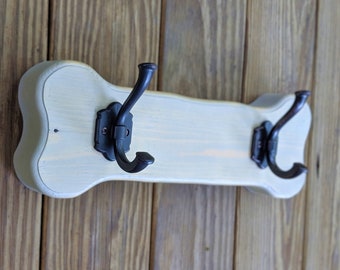 Può includere: Un attaccapanni in legno a forma di osso, dipinto di color crema chiaro, con due ganci in metallo nero. L'attaccapanni è montato su una parete di legno con assi verticali.