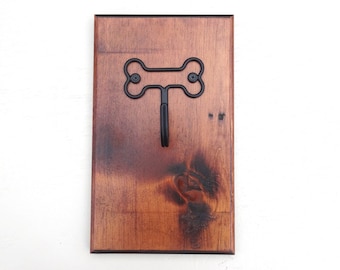 Può includere: Un gancio da parete in legno con finitura marrone scuro. Il gancio presenta un design a forma di osso in metallo nero con un singolo gancio sottostante. La venatura del legno è visibile e il gancio è montato su una tavola di legno rettangolare.