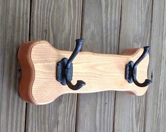 Può includere: Appendiabiti in legno chiaro con due ganci in metallo nero. L'appendiabiti ha una forma curva unica ed è montato su una parete in legno. I ganci sono ornati e offrono un'estetica rustica.