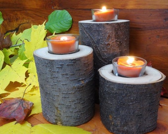 Può includere: Tre portacandele rustici in legno con candele arancioni accese in contenitori di vetro. I portacandele sono realizzati con sezioni di tronchi d'albero, che mostrano la naturale consistenza della corteccia. Foglie autunnali gialle, verdi e rosse circondano i portacandele.