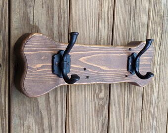 Può includere: Un attaccapanni in legno con finitura marrone scuro e due ganci in metallo nero. L'attaccapanni ha una forma curva decorativa ed è montato su una parete di legno. I ganci sono ornati.