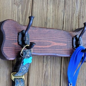 以下が含まれることがあります： 3つの黒い金属製のフックが付いた、犬の骨の形をした木製の壁掛けフック。青い犬の首輪と青いリードがフックに掛けられています。木材は濃い茶色に着色されており、木製の壁に取り付けられています。