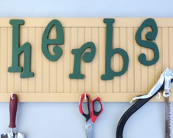 Può includere: Un cartello di legno giallo con la scritta "herbs" in verde. Sotto, vari attrezzi da giardinaggio sono appesi a ganci, tra cui una cazzuola, forbici e una sega.