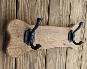 Può includere: Appendiabiti in legno con due ganci in metallo nero. Il portabiti è realizzato in legno marrone chiaro e invecchiato ed è montato su una parete di legno. I ganci hanno un design curvo e sono fissati con placche nere decorative.