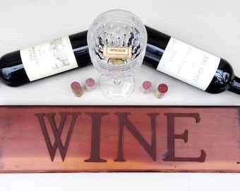 Può includere: Un'insegna in legno con la scritta "WINE" intagliata, insieme a due bottiglie di vino rosso, un calice di cristallo e diversi tappi. Le bottiglie di vino sono nere con etichette bianche e lamina rossa.