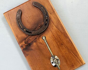 Può includere: Un attaccapanni da parete in legno con un ferro di cavallo e un gancio in ottone. La tavola rettangolare in legno è tinta di un caldo colore marrone. Il ferro di cavallo è marrone scuro e il gancio è color ottone.
