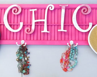 Può includere: Un organizer da parete rosa con la scritta "CHIC" in caratteri bianchi. L'organizer ha ganci che sostengono un berretto blu, collane e un cappello di paglia. Lo sfondo è una parete grigio chiaro.