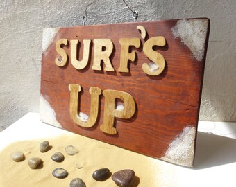 May include: Wooden sign with the words "SURF'S UP" in light brown letters. The sign is a reddish-brown color with white accents on the corners. Small stones and sand are in the foreground.